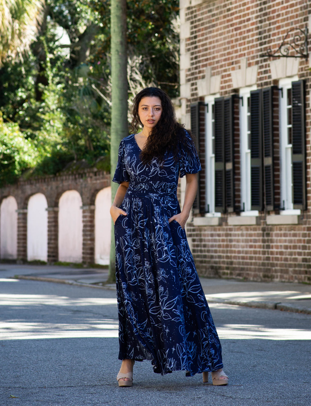 Woman in a blue dress standing on a street with palm trees and a brick building in the background